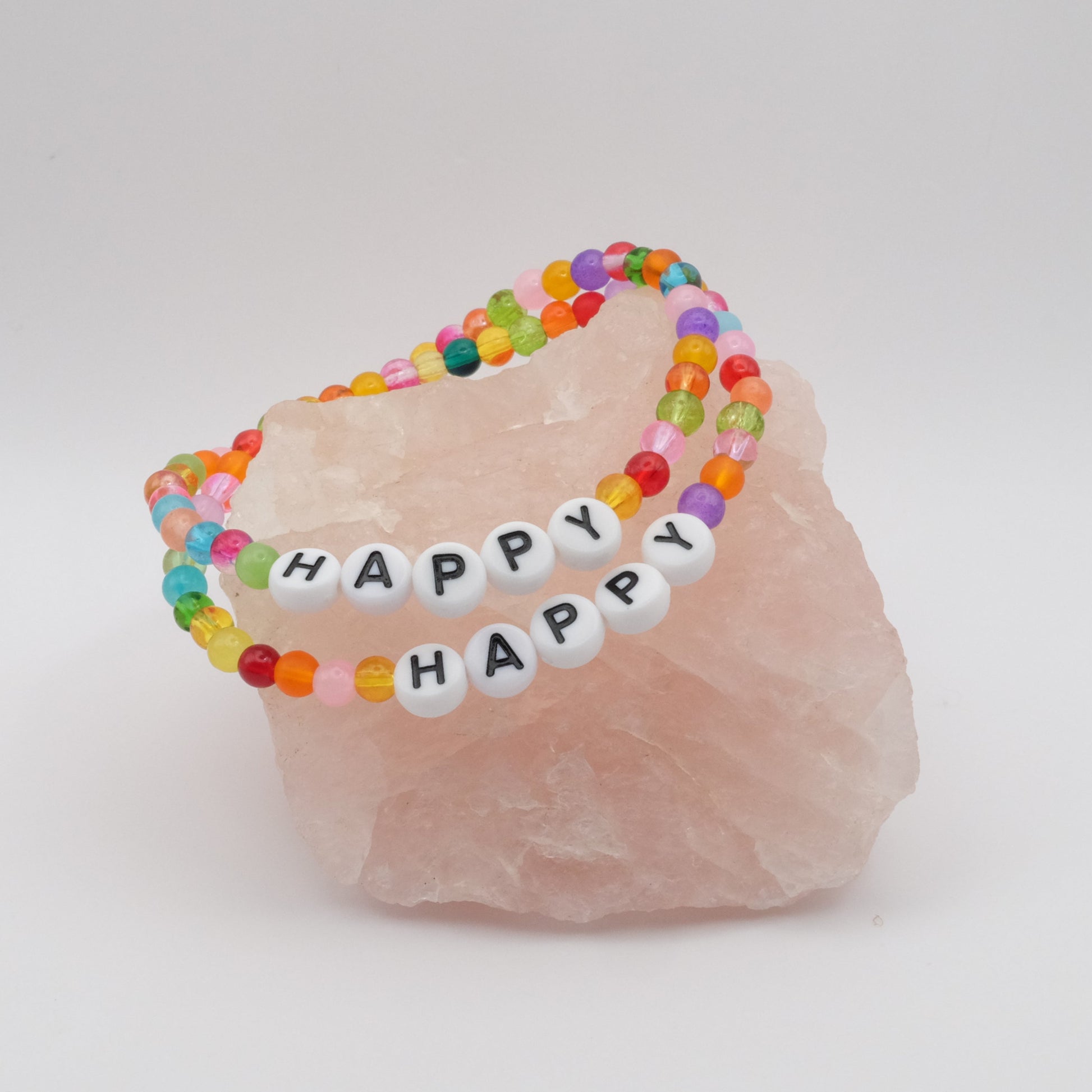 Two elastic beaded bracelets composed of multicoloured 4mm glass beads with white acrylic letter beads spelling “HAPPY”, displayed on a pale pink crystal rock.