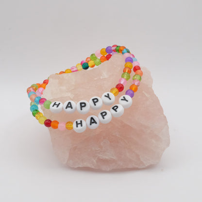 Two elastic beaded bracelets composed of multicoloured 4mm glass beads with white acrylic letter beads spelling “HAPPY”, displayed on a pale pink crystal rock.