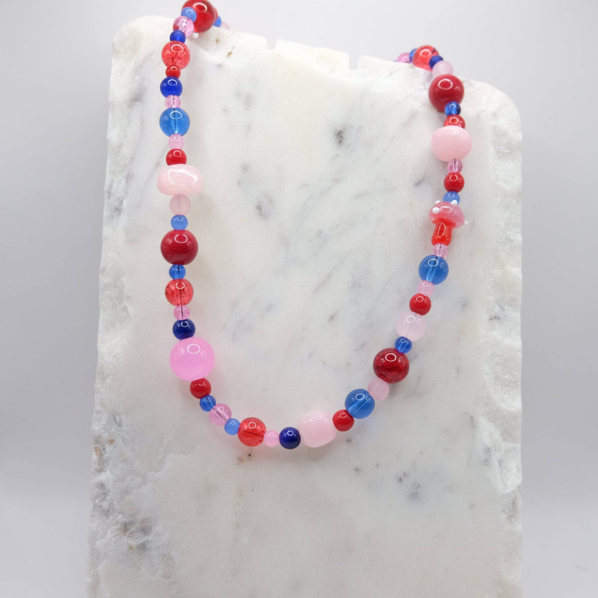 A beaded necklace composed of mixed-size pink, red and blue glass beads and pink dyed jade—featuring a small blown glass toadstool bead, shown on a white marble background.
