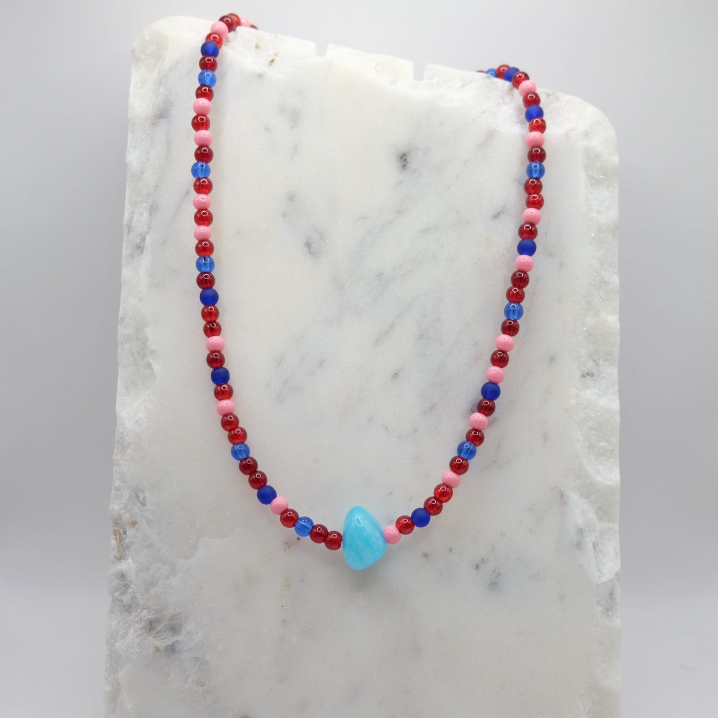 A beaded necklace composed of red, pink and blue glass beads - featuring a central sky-blue bead, shown on a white marble background.