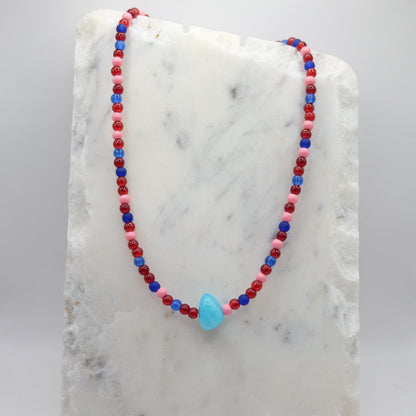 A beaded necklace composed of red, pink and blue glass beads - featuring a central sky-blue bead, shown on a white marble background.