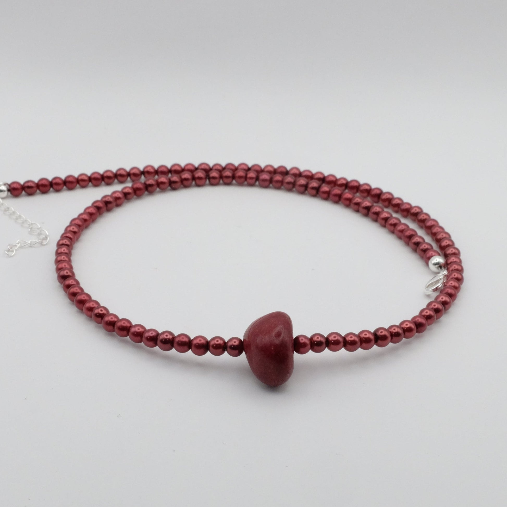 A handmade beaded necklace featuring metallic burnt red 4mm glass beads and a smooth red dyed jade focal bead, displayed on a white marble stand.
