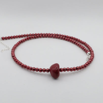 A handmade beaded necklace featuring metallic burnt red 4mm glass beads and a smooth red dyed jade focal bead, displayed on a white marble stand.