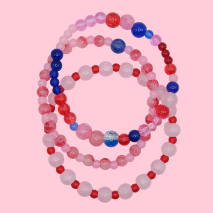 A one of a kind bracelet stack composed of layered glass bead bracelets in bold pink, red and blue tones, shown on a soft neutral surface.
