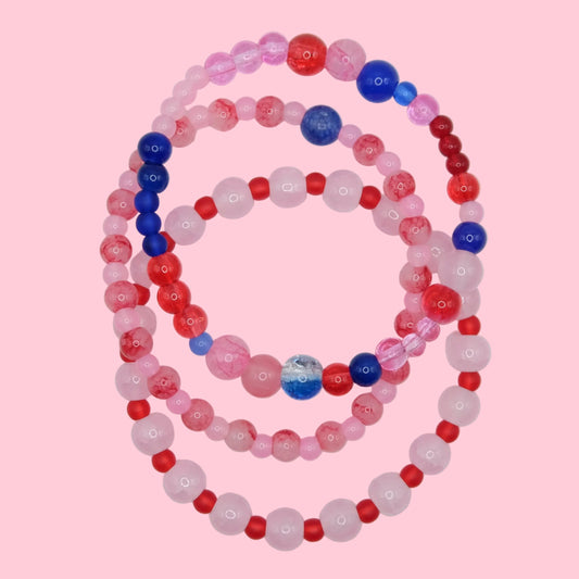 A one of a kind bracelet stack composed of layered glass bead bracelets in bold pink, red and blue tones, shown on a soft neutral surface.