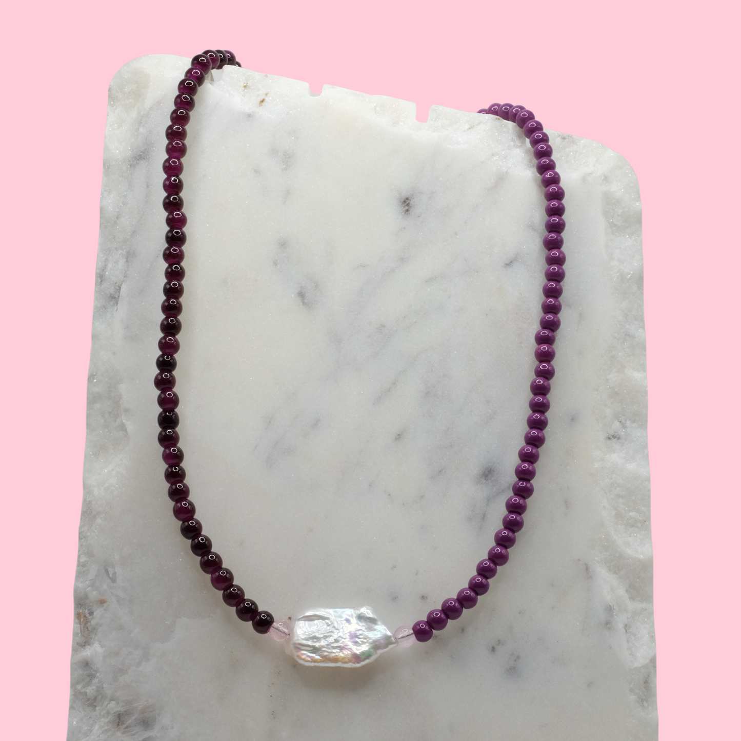 Purple beaded necklace with a clear crystal pendant on a marble surface with a pink background