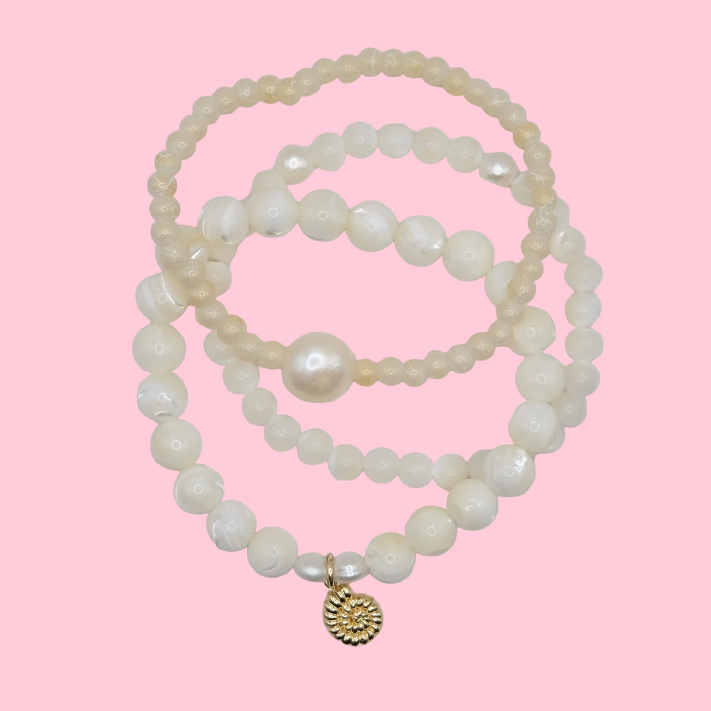 Stack of mother of pearl and pearl bracelets with a gold nautilus seashell charm on a pink background