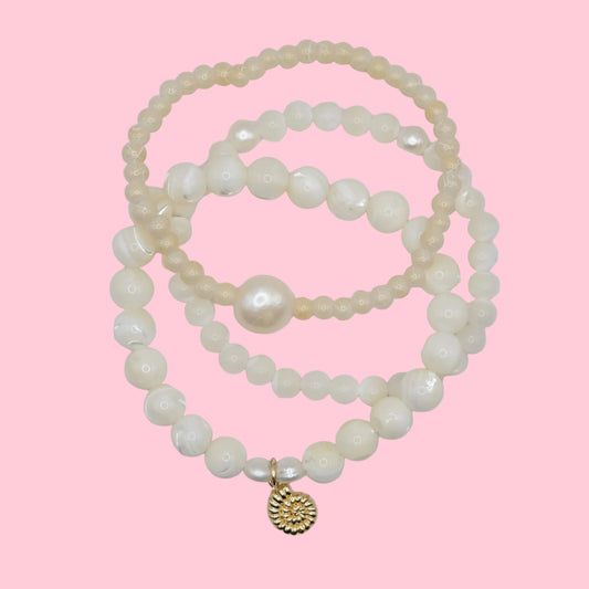 Stack of mother of pearl and pearl bracelets with a gold nautilus seashell charm on a pink background