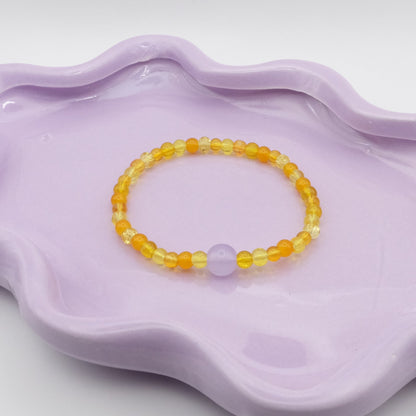 An elastic beaded bracelet made from 4mm glass beads in a mix of yellow glass beads, accented by a single 8mm lavender feature bead, arranged on a wavy lavender ceramic dish with a clean background.