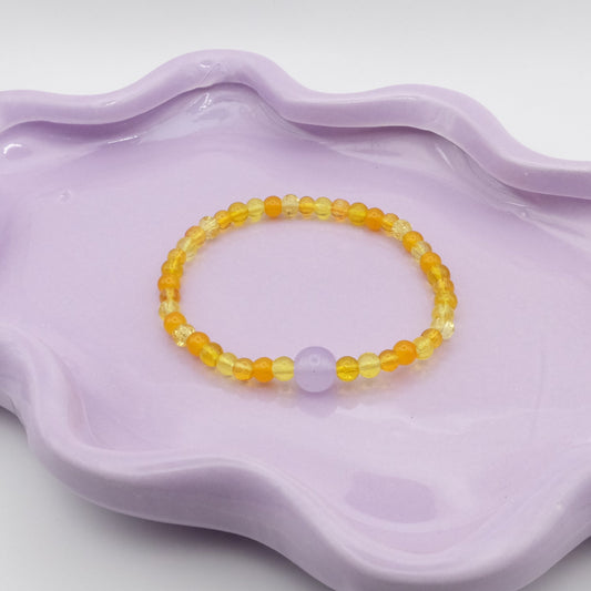 An elastic beaded bracelet made from 4mm glass beads in a mix of yellow glass beads, accented by a single 8mm lavender feature bead, arranged on a wavy lavender ceramic dish with a clean background.