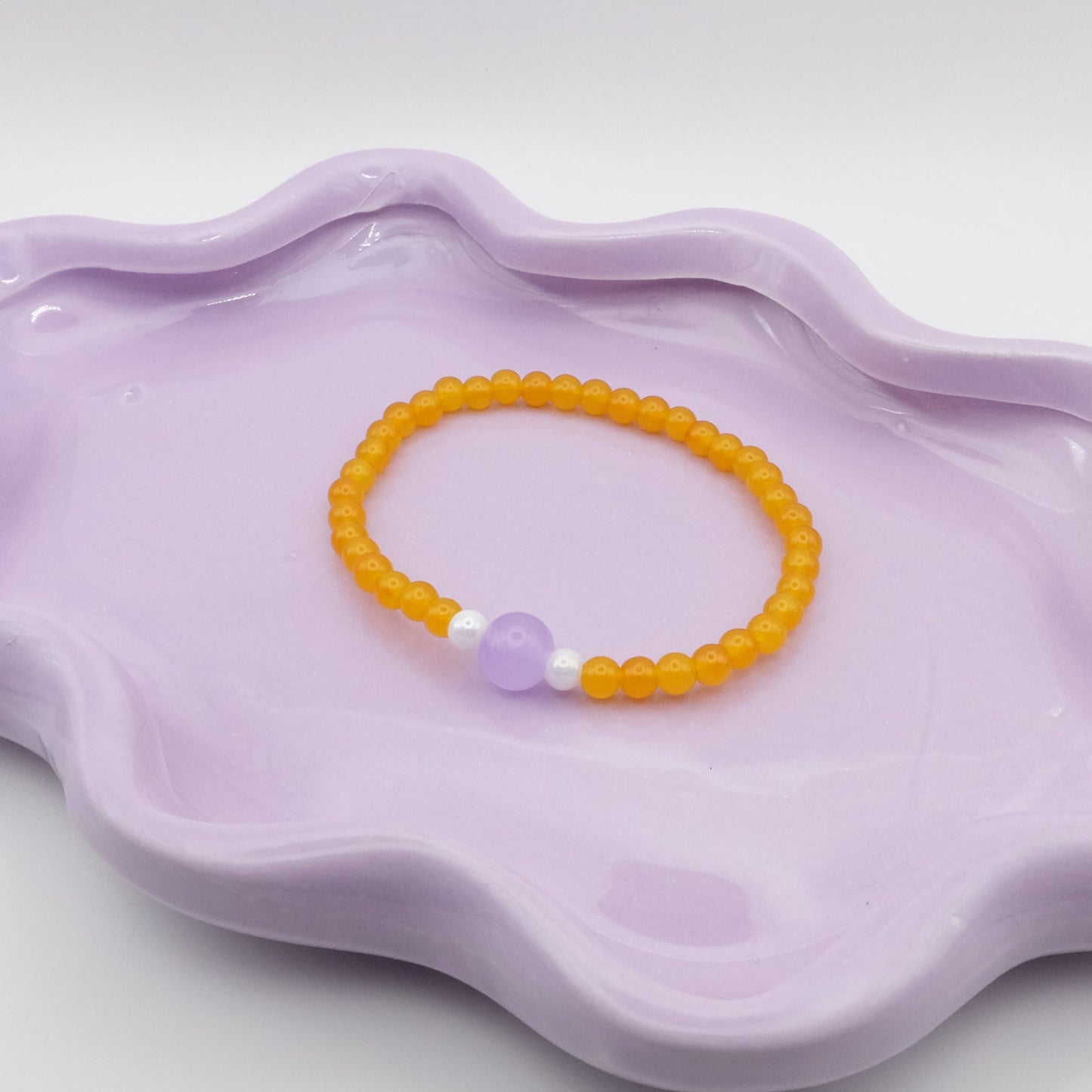 An elastic beaded bracelet crafted from 4mm sunny yellow glass beads with a single 8mm lavender feature bead, flanked by two 4mm white metallic beads, arranged on a wavy lavender ceramic dish with a simple background.