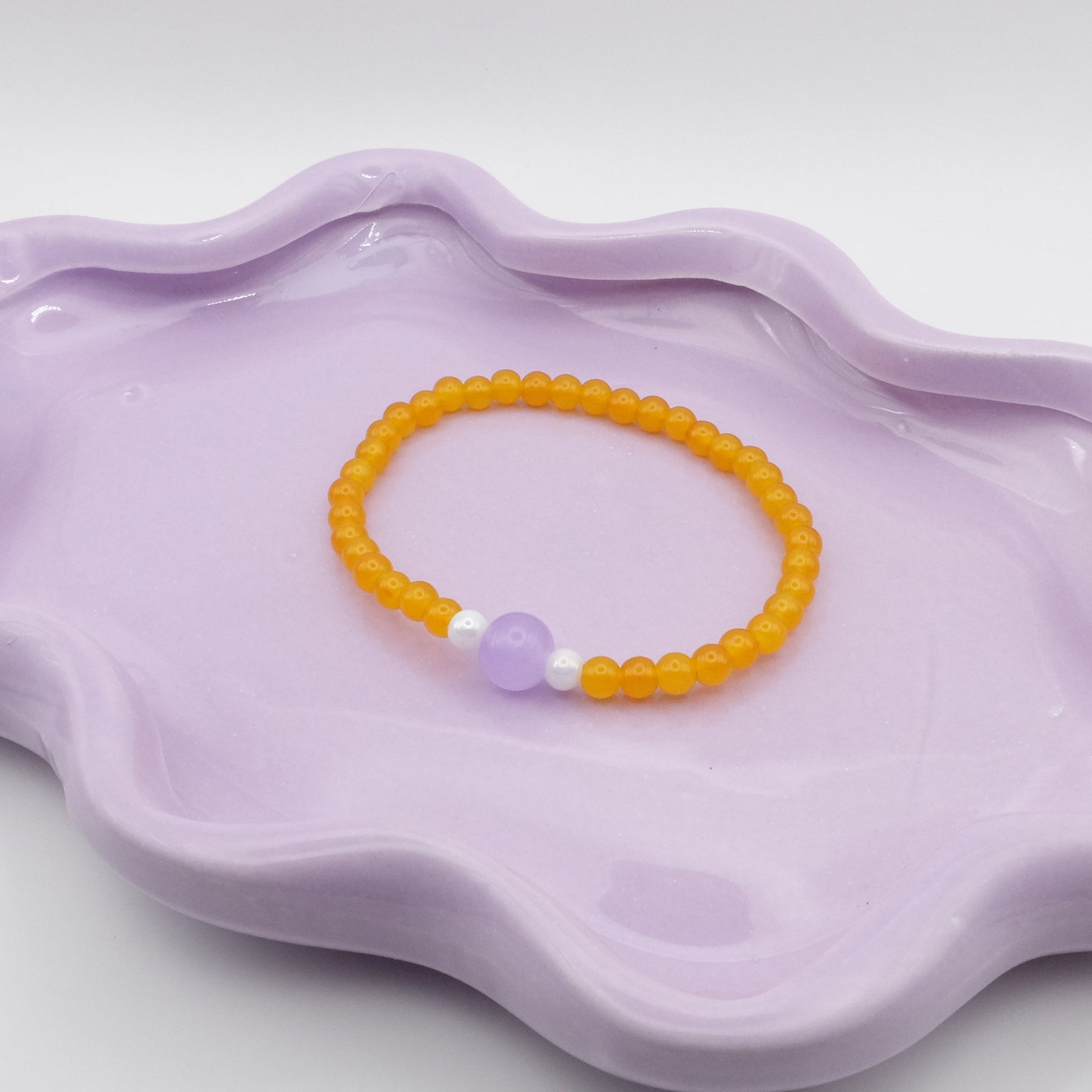An elastic beaded bracelet crafted from 4mm sunny yellow glass beads with a single 8mm lavender feature bead, flanked by two 4mm white metallic beads, arranged on a wavy lavender ceramic dish with a simple background.