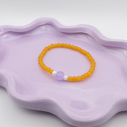 An elastic beaded bracelet crafted from 4mm sunny yellow glass beads with a single 8mm lavender feature bead, flanked by two 4mm white metallic beads, arranged on a wavy lavender ceramic dish with a simple background.