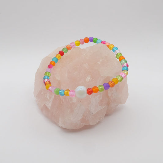 An elastic beaded bracelet composed of red, orange, yellow,  green, aqua, blue, purple and pink 4mm glass beads in a random pattern with a single white pearlescent 8mm feature bead, displayed on a pale pink crystal rock against a soft white background.