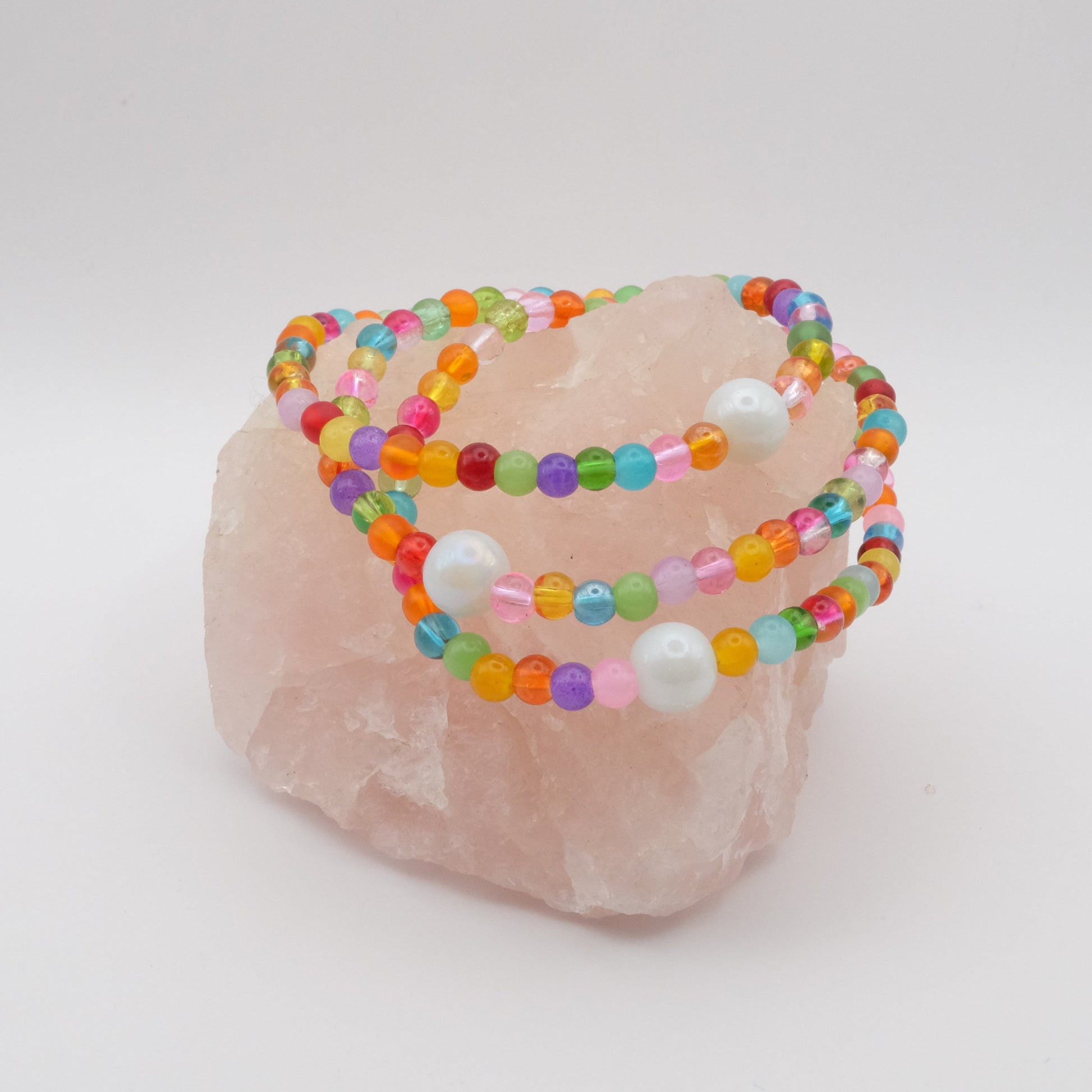 A stack of three elastic beaded bracelets composed of red, orange, yellow,  green, aqua, blue, purple and pink 4mm glass beads in a random pattern with a single white pearlescent 8mm feature bead, displayed on a pale pink crystal rock against a soft white background.