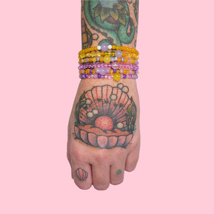 A tattooed arm with a vibrant octopus and seashell design is shown wearing multiple stacked elastic beaded bracelets in bright yellow and purple hues, set against a solid pink background.