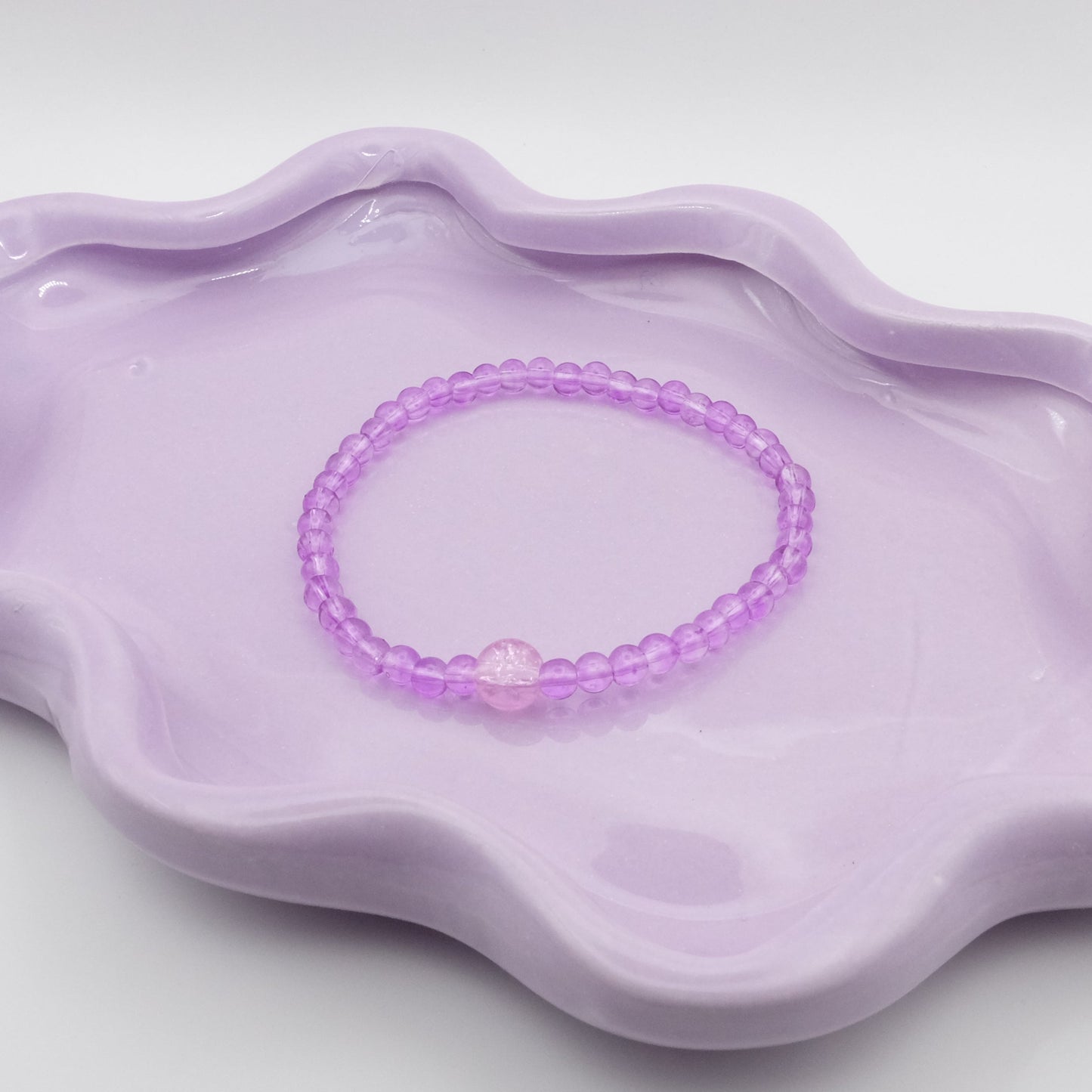 An elastic beaded bracelet made entirely from 4mm transparent purple glass beads, featuring a single 8mm crackle purple bead in the center, arranged on a wavy lavender ceramic dish with a simple background.