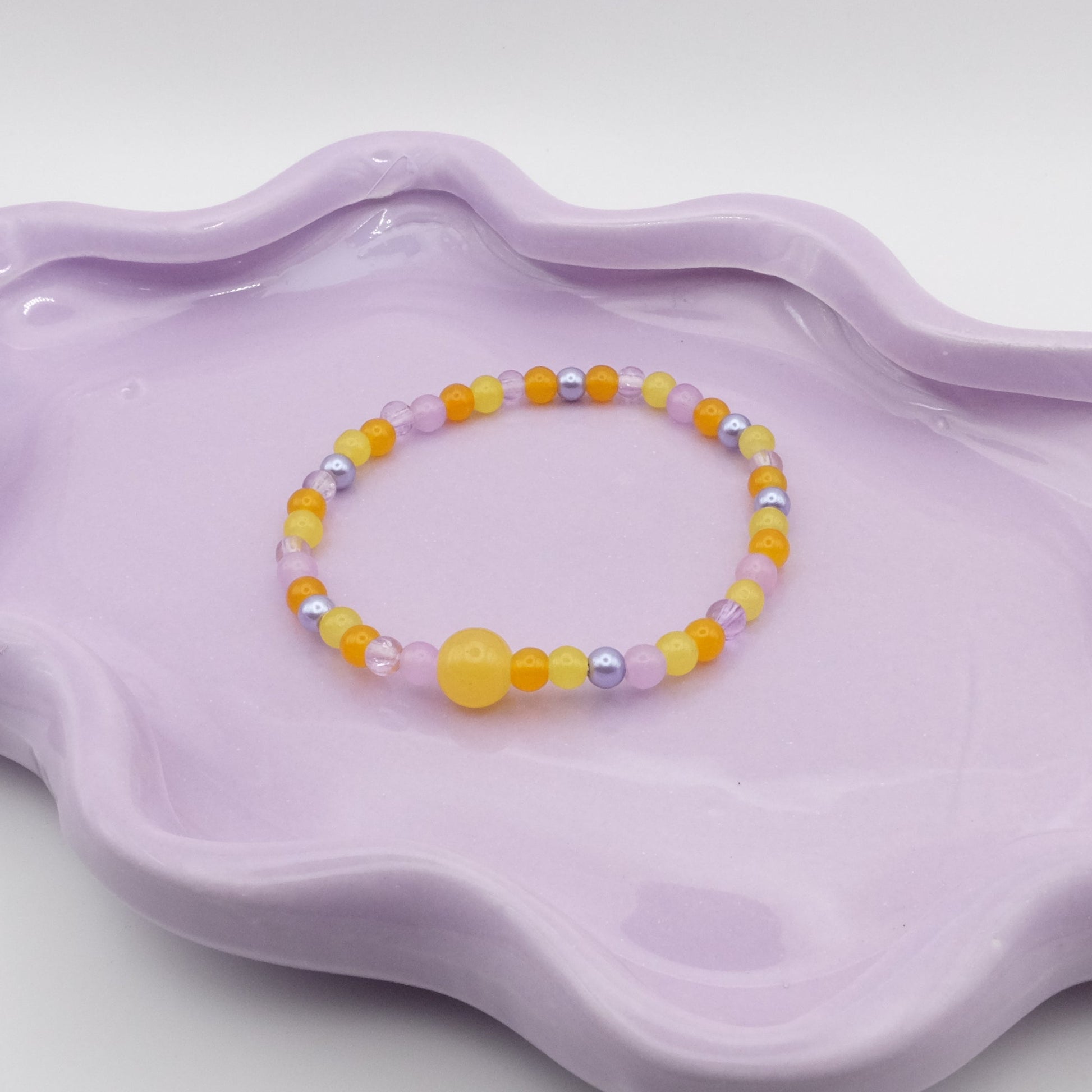 An elastic beaded bracelet featuring a mix of 4mm yellow and purple glass beads, highlighted by a single 8mm yellow feature bead, arranged on a wavy lavender ceramic dish with a simple background.