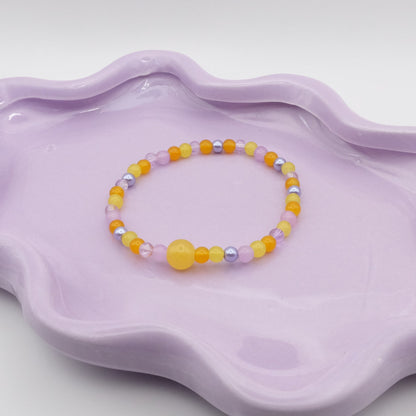 An elastic beaded bracelet featuring a mix of 4mm yellow and purple glass beads, highlighted by a single 8mm yellow feature bead, arranged on a wavy lavender ceramic dish with a simple background.