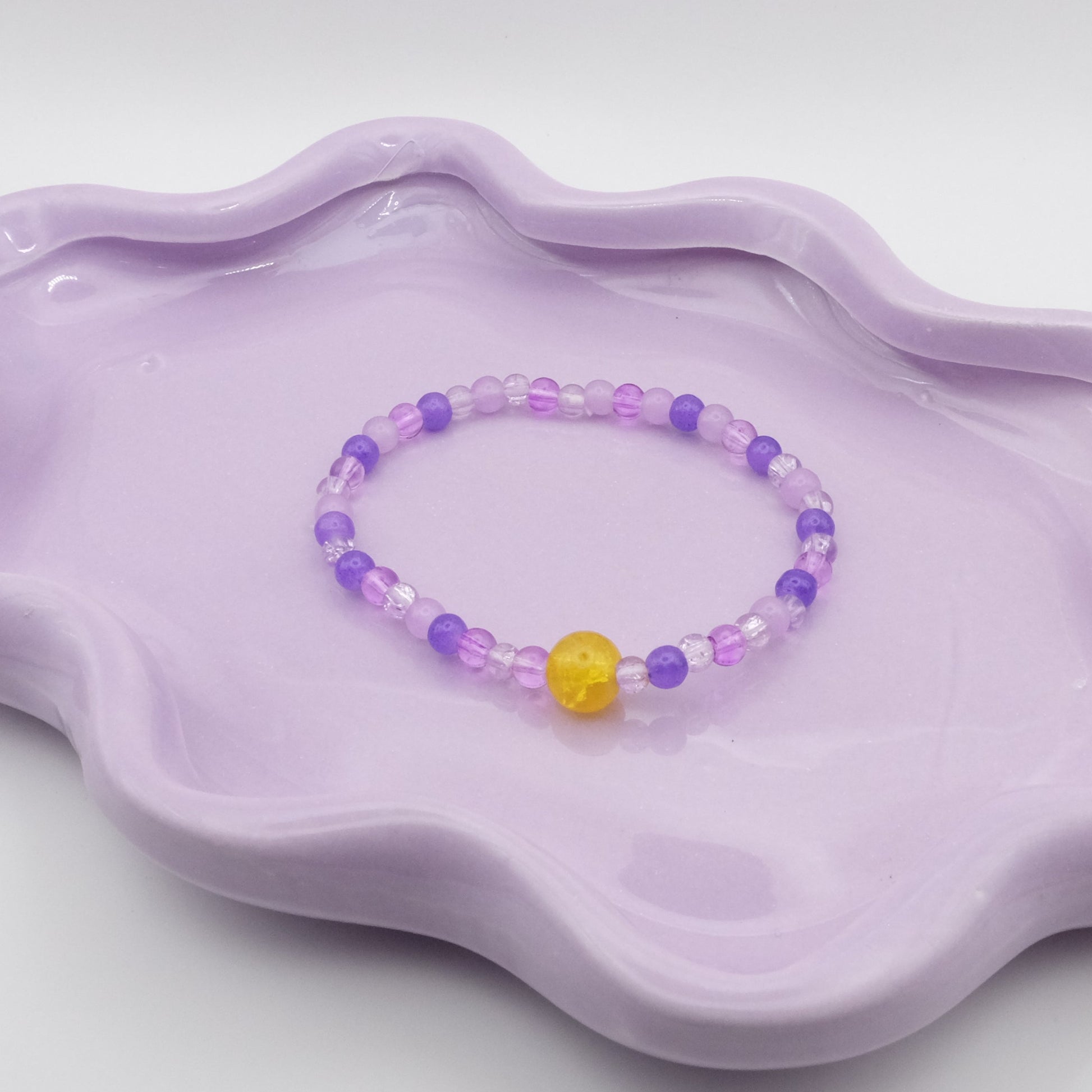An elastic beaded bracelet composed of 4mm glass beads in a mix of purples, accented by a single 8mm yellow feature bead, arranged on a wavy lavender ceramic dish with a simple background.
