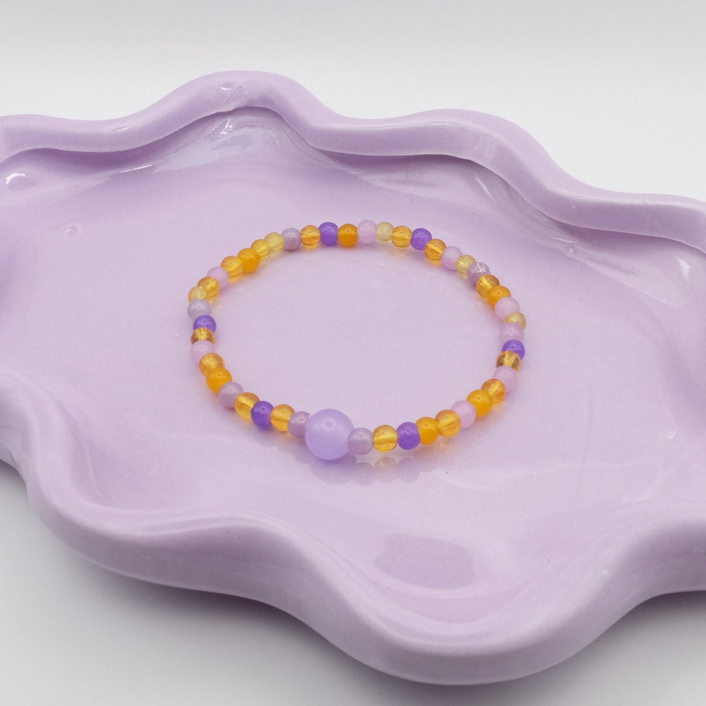 An elastic beaded bracelet combining 4mm purple and yellow glass beads in various shades, accented by a single 8mm purple feature bead, arranged on a wavy lavender ceramic dish with a simple background.