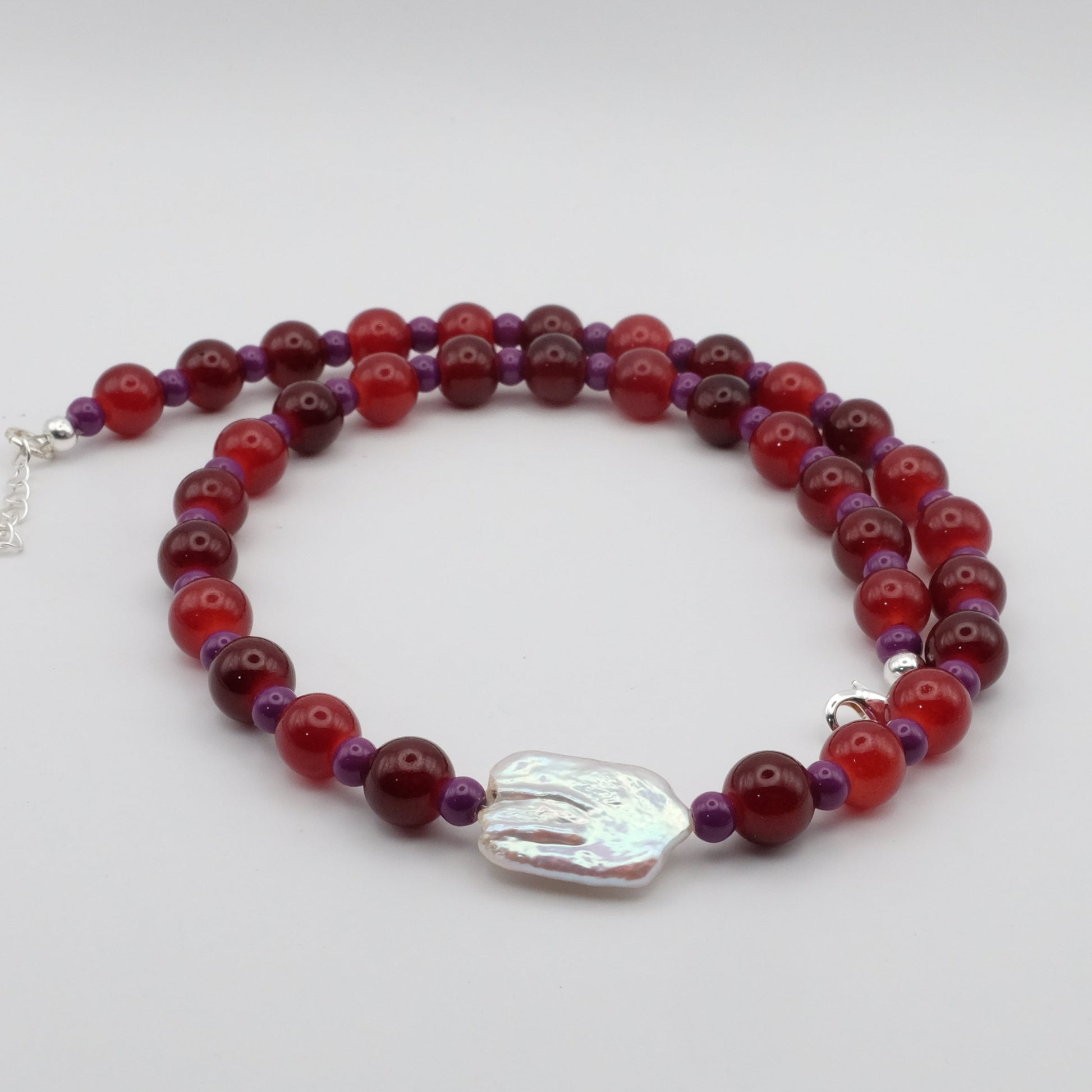A handmade beaded necklace featuring alternating 8mm red glass beads and 4mm deep purple glass beads with a white baroque freshwater pearl centrepiece, displayed on a white background.