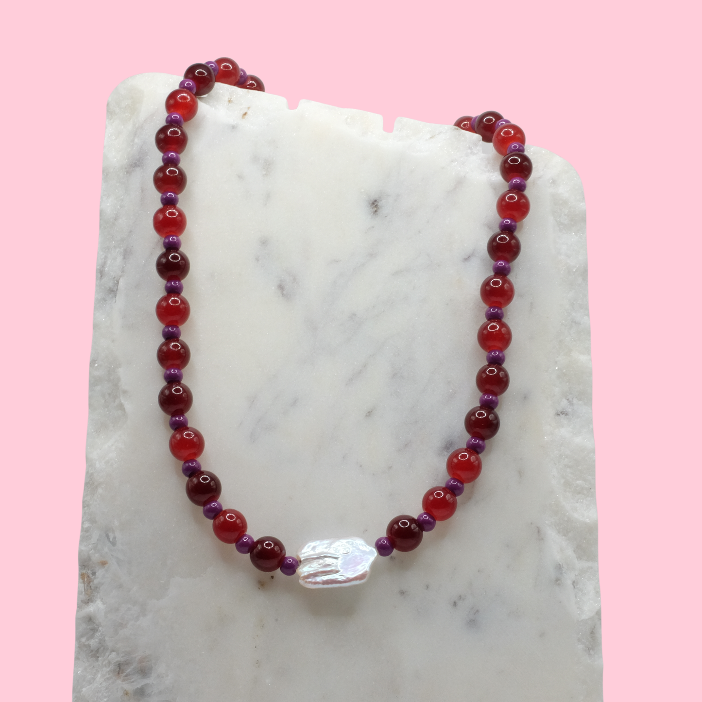 Beaded necklace with red and purple beads on a marble surface with a pink background