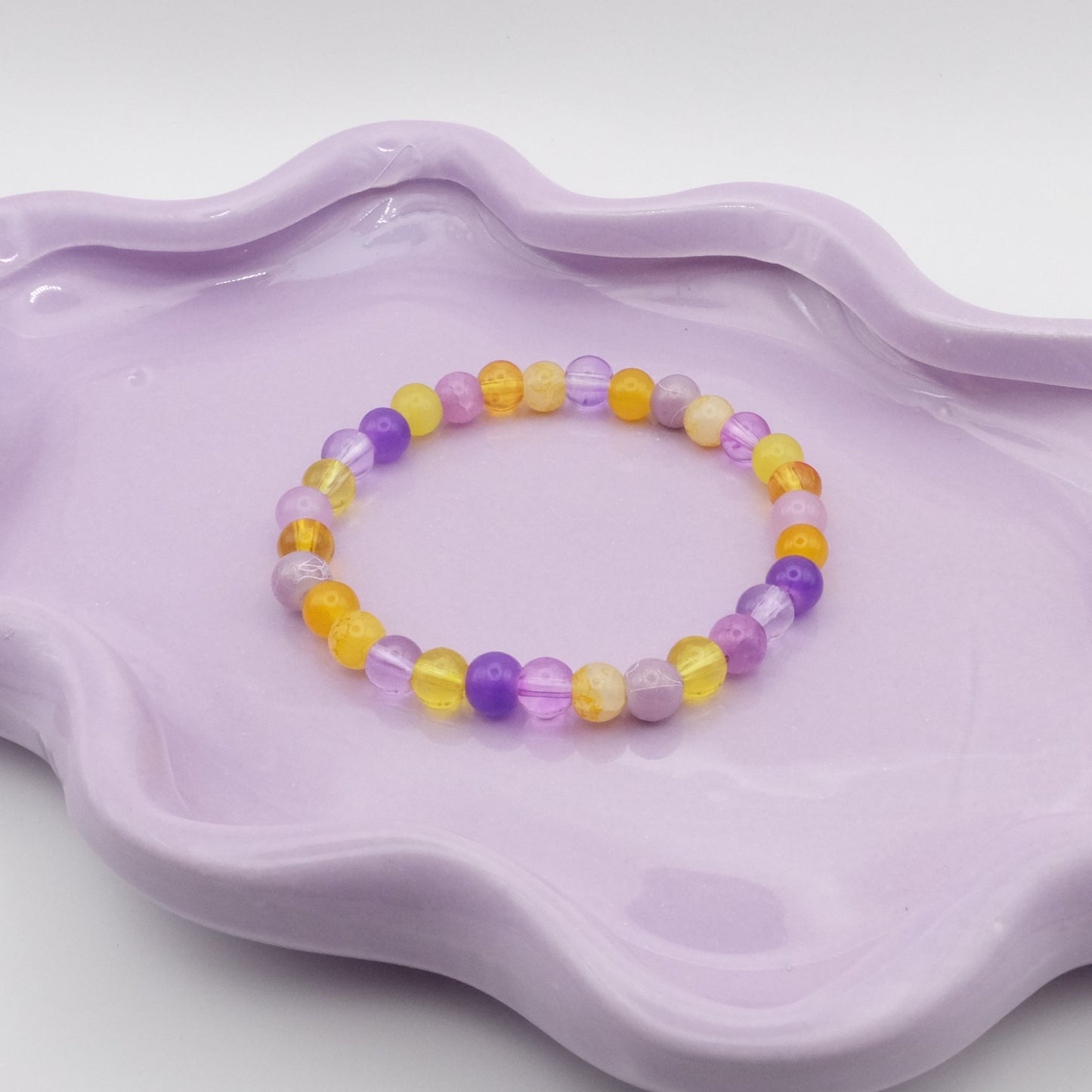 An elastic beaded bracelet made from 6mm glass beads in a moody-meets-mellow blend of light and dark purples with touches of yellow, shown on a wavy lavender dish before a clean, minimal background.