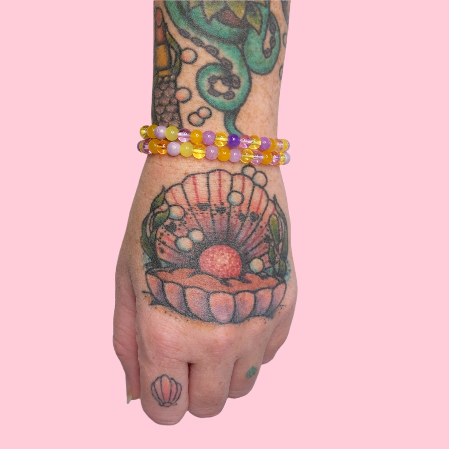 A tattooed arm featuring an octopus and seashell design is shown wearing multiple elastic beaded bracelets in purple and yellow hues, set against a solid pink background.