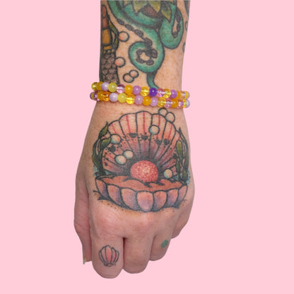 A tattooed arm featuring an octopus and seashell design is shown wearing multiple elastic beaded bracelets in purple and yellow hues, set against a solid pink background.