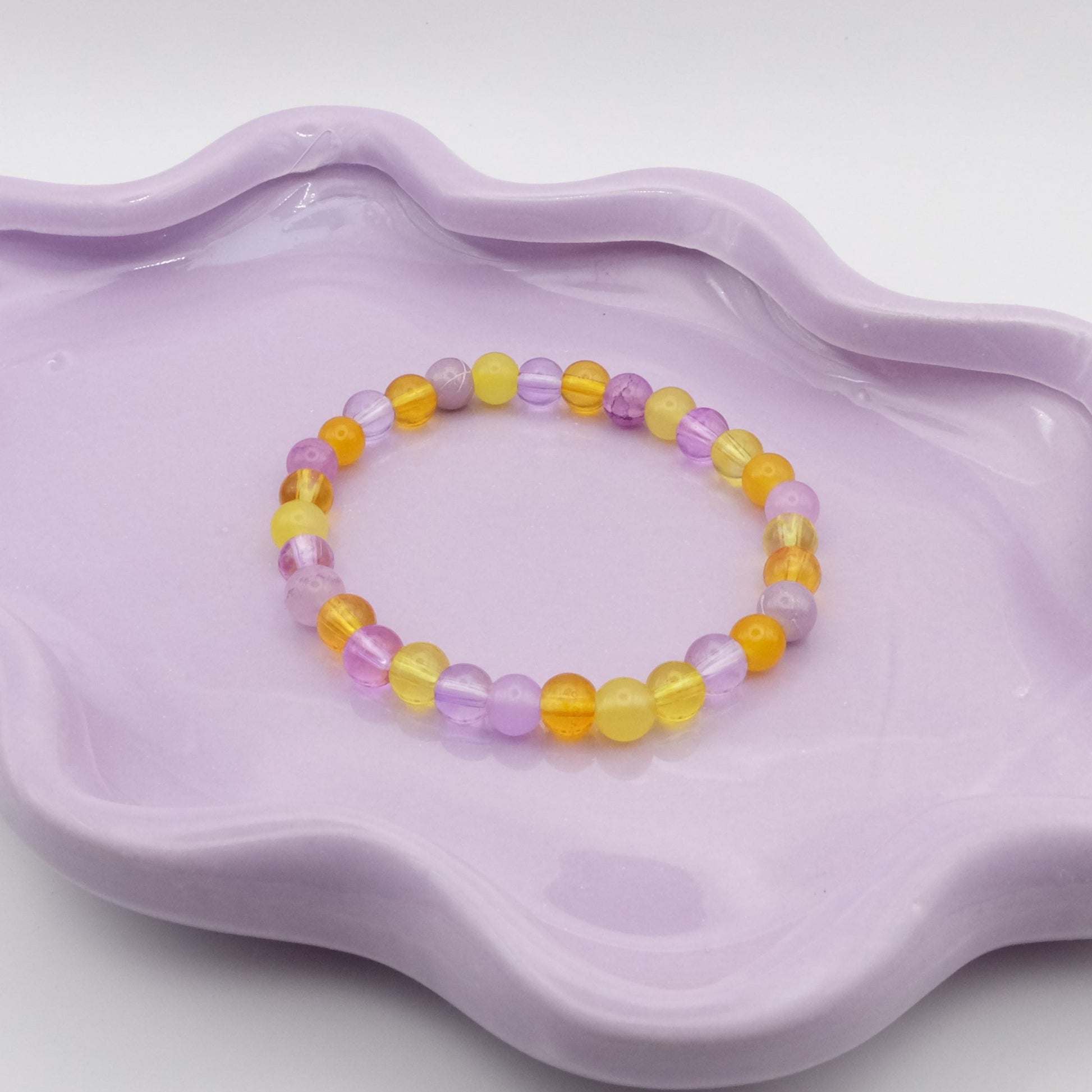 An elastic beaded bracelet crafted entirely from 6mm glass beads in a cheerful mix of light purples and sunny yellows, displayed on a wavy lavender dish against a simple, light backdrop.