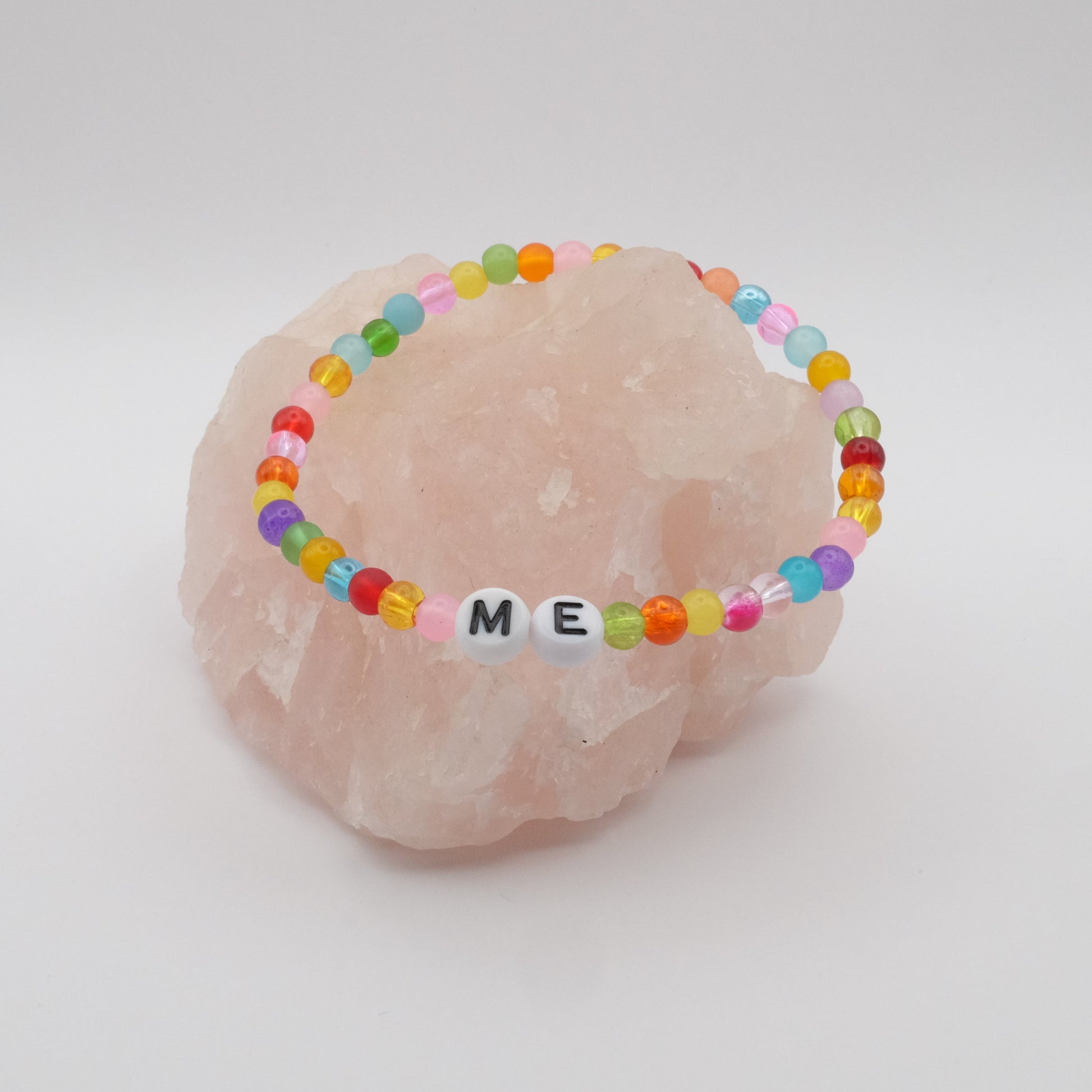 An elastic beaded bracelet composed of colourful 4mm glass beads in red, orange, yellow, green, aqua, blue, purple and pink with white acrylic letter beads spelling “ME,” displayed on a pale pink crystal rock.