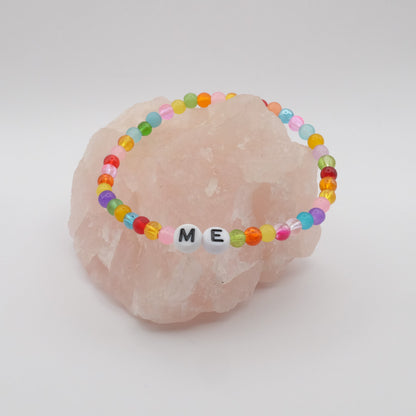 An elastic beaded bracelet composed of colourful 4mm glass beads in red, orange, yellow, green, aqua, blue, purple and pink with white acrylic letter beads spelling “ME,” displayed on a pale pink crystal rock.