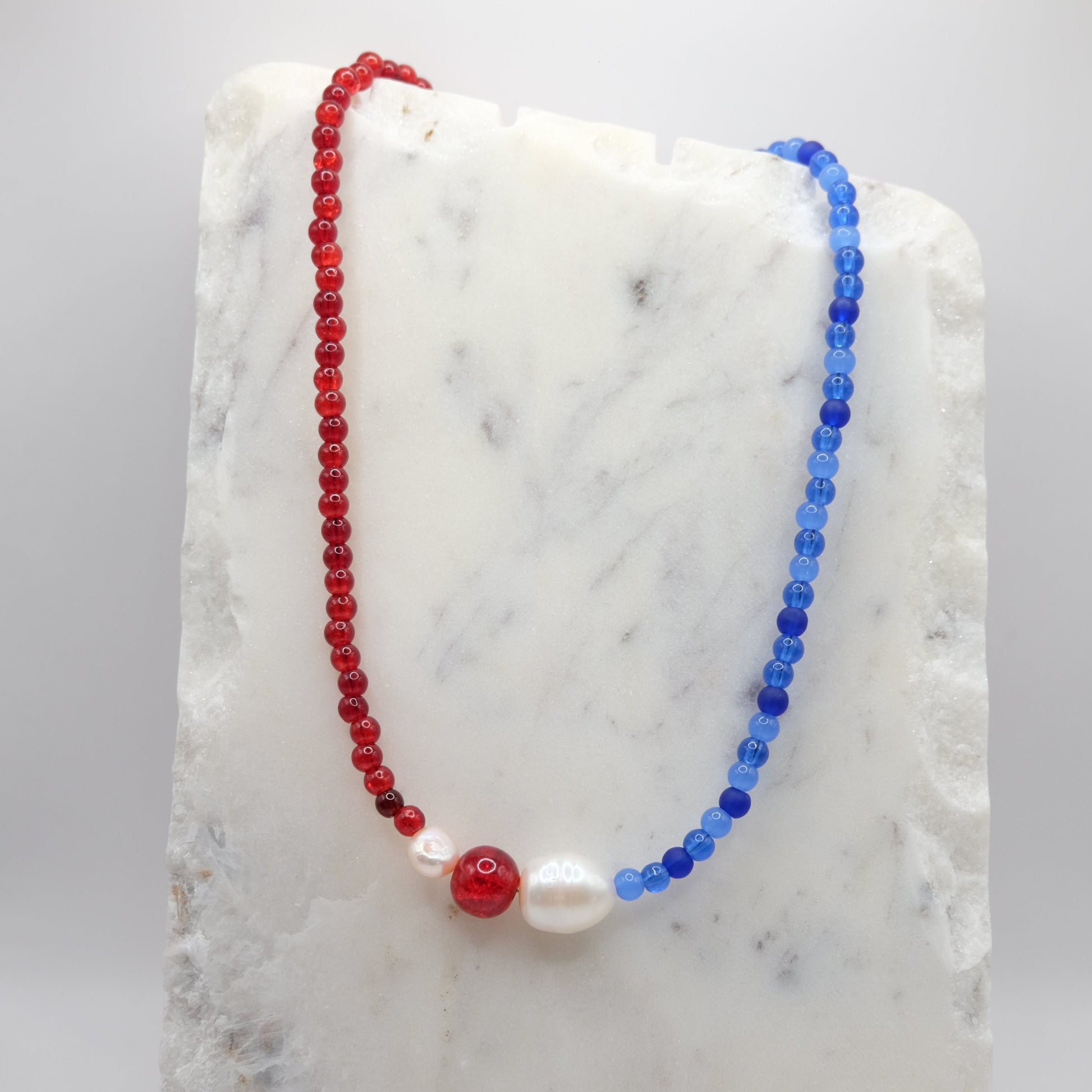 A beaded necklace composed of red and blue glass beads - featuring a freshwater pearl nugget, red glitter bead and pink pearl, shown on a white marble background.