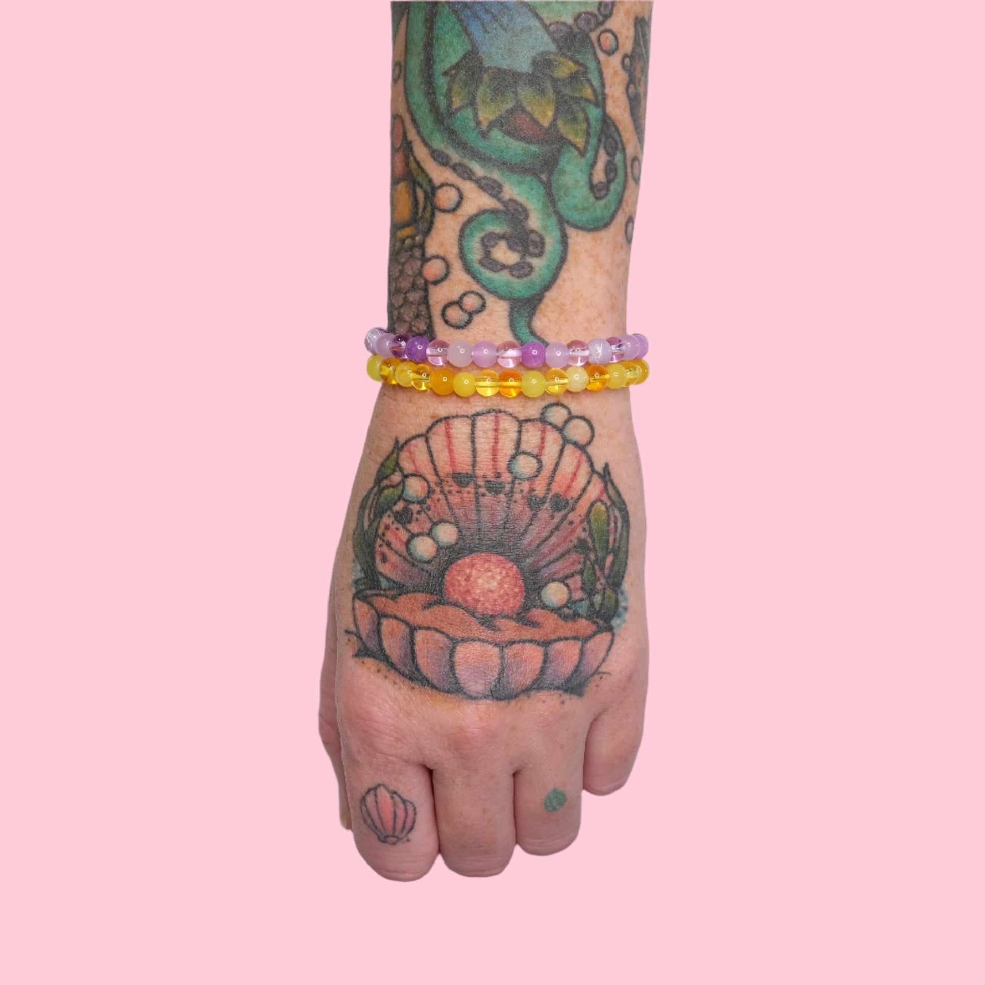 An arm adorned with colorful octopus and seashell tattoos wears two elastic beaded bracelets in lavender and sunny yellow tones, set against a solid pink background.