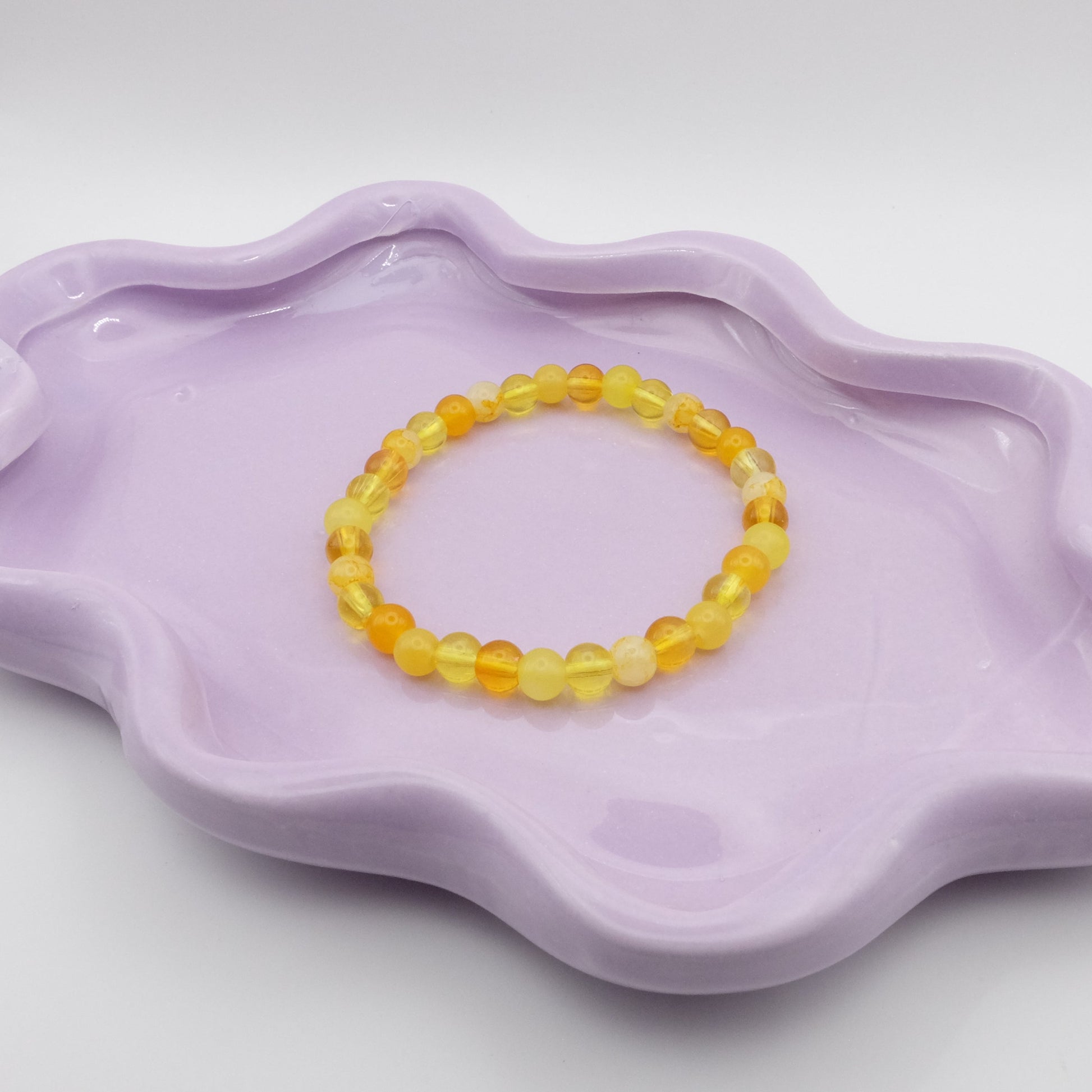 An elastic beaded bracelet crafted entirely from 6mm yellow glass beads in a random mix of shades and finishes including solid, crackle, and frosted. Displayed on a wavy lavender dish against a simple, light backdrop.