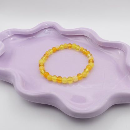 An elastic beaded bracelet crafted entirely from 6mm yellow glass beads in a random mix of shades and finishes including solid, crackle, and frosted. Displayed on a wavy lavender dish against a simple, light backdrop.