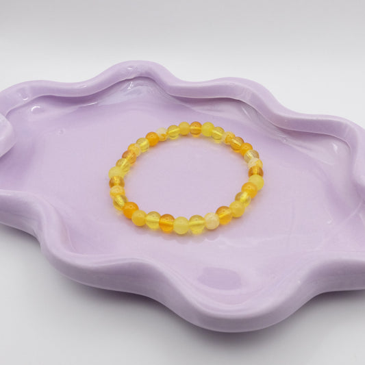An elastic beaded bracelet crafted entirely from 6mm yellow glass beads in a random mix of shades and finishes including solid, crackle, and frosted. Displayed on a wavy lavender dish against a simple, light backdrop.