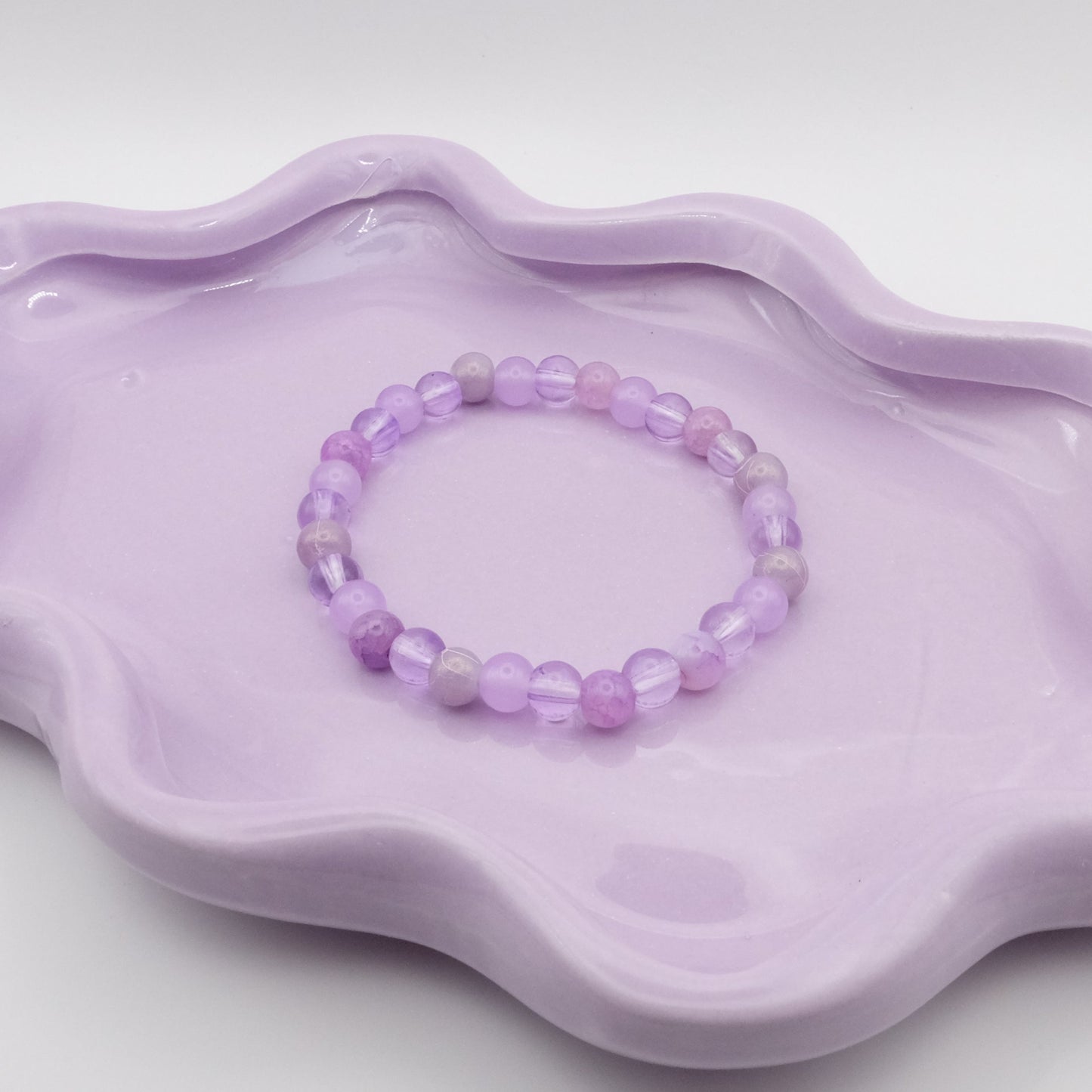 An elastic beaded bracelet crafted entirely from 6mm purple glass beads in a random mix of shades and finishes including solid, crackle, and frosted. Displayed on a wavy lavender dish against a simple, light backdrop.