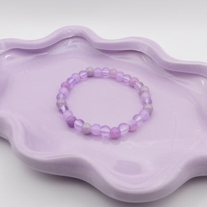An elastic beaded bracelet crafted entirely from 6mm purple glass beads in a random mix of shades and finishes including solid, crackle, and frosted. Displayed on a wavy lavender dish against a simple, light backdrop.