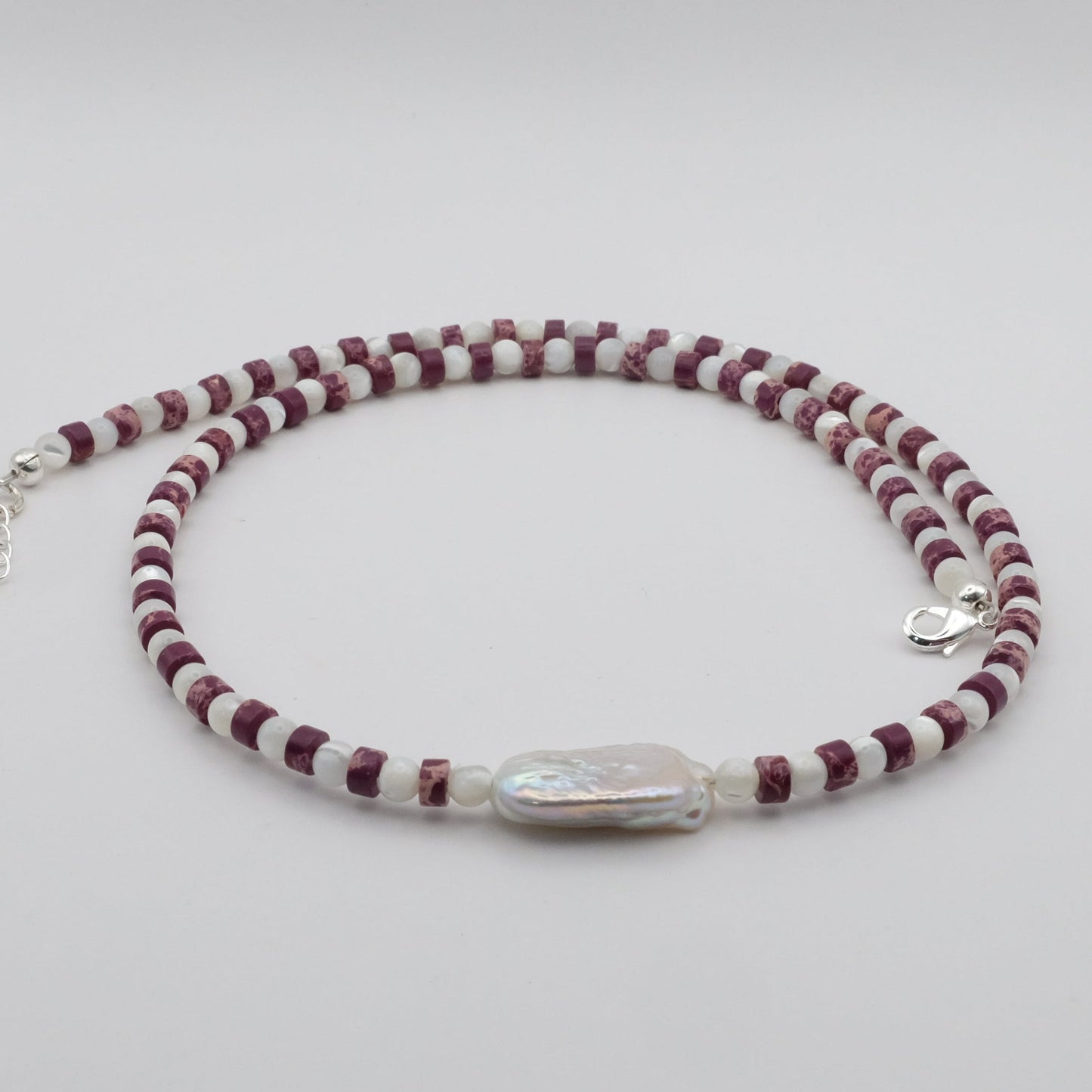 A handmade beaded necklace featuring purple sea sediment jasper heishi gemstone beads, mother of pearl beads and a baroque freshwater pearl focal bead, displayed on a white background.