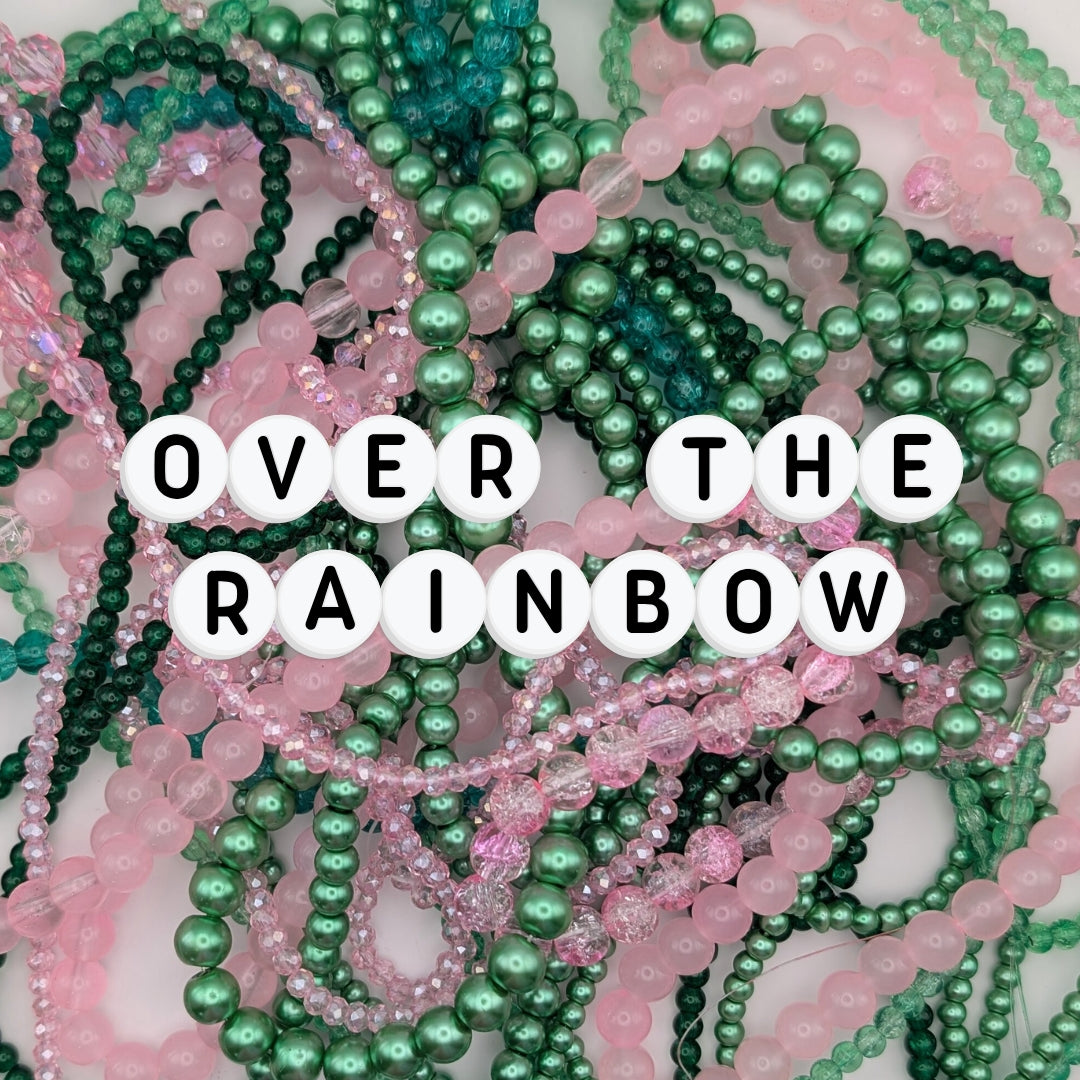 A dreamy pile of pink and green elastic beaded bracelets made from frosted, glossy, and faceted glass beads, with acrylic letter beads spelling “OVER THE RAINBOW.” A whimsical handmade jewellery collection perfect for dopamine dressing, quirky jewellery, and colourful accessories