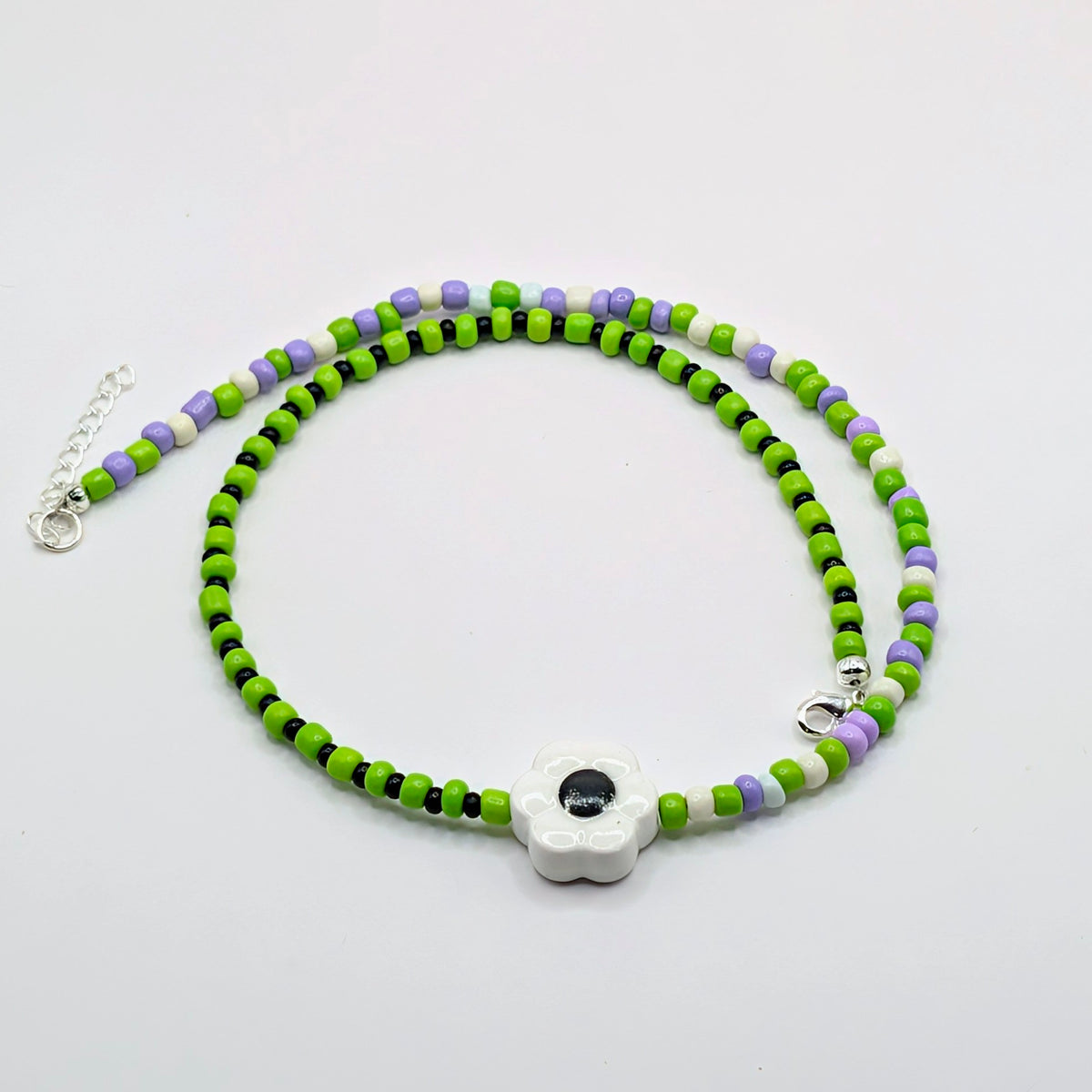 A beaded necklace featuring bright green, lime green, white, and lavender purple beads, with a central white flower charm containing a black evil eye. The necklace is displayed on a clean white background with its silver clasp and extender chain visible.