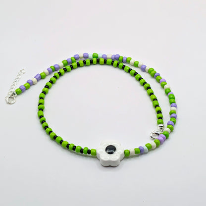 A beaded necklace featuring bright green, lime green, white, and lavender purple beads, with a central white flower charm containing a black evil eye. The necklace is displayed on a clean white background with its silver clasp and extender chain visible.