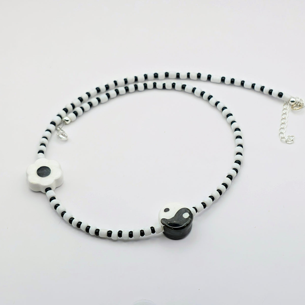 A beaded necklace with alternating black and white beads, featuring a white flower-shaped bead with a black center, a black and white yin-yang charm, and a silver-tone clasp with extender chain.
