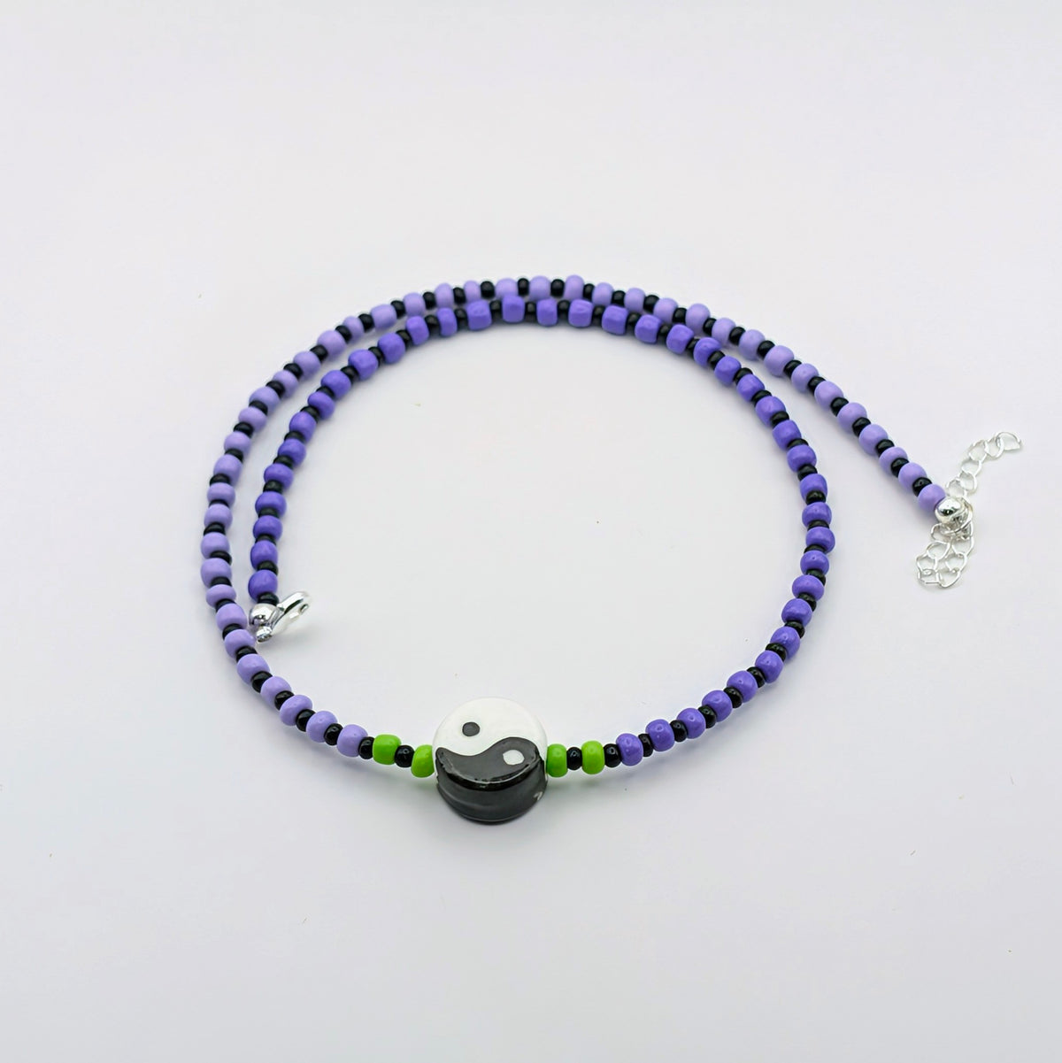 A beaded necklace with alternating purple and black beads, accented by small green beads and a central black and white yin-yang charm, displayed on a clean white background.