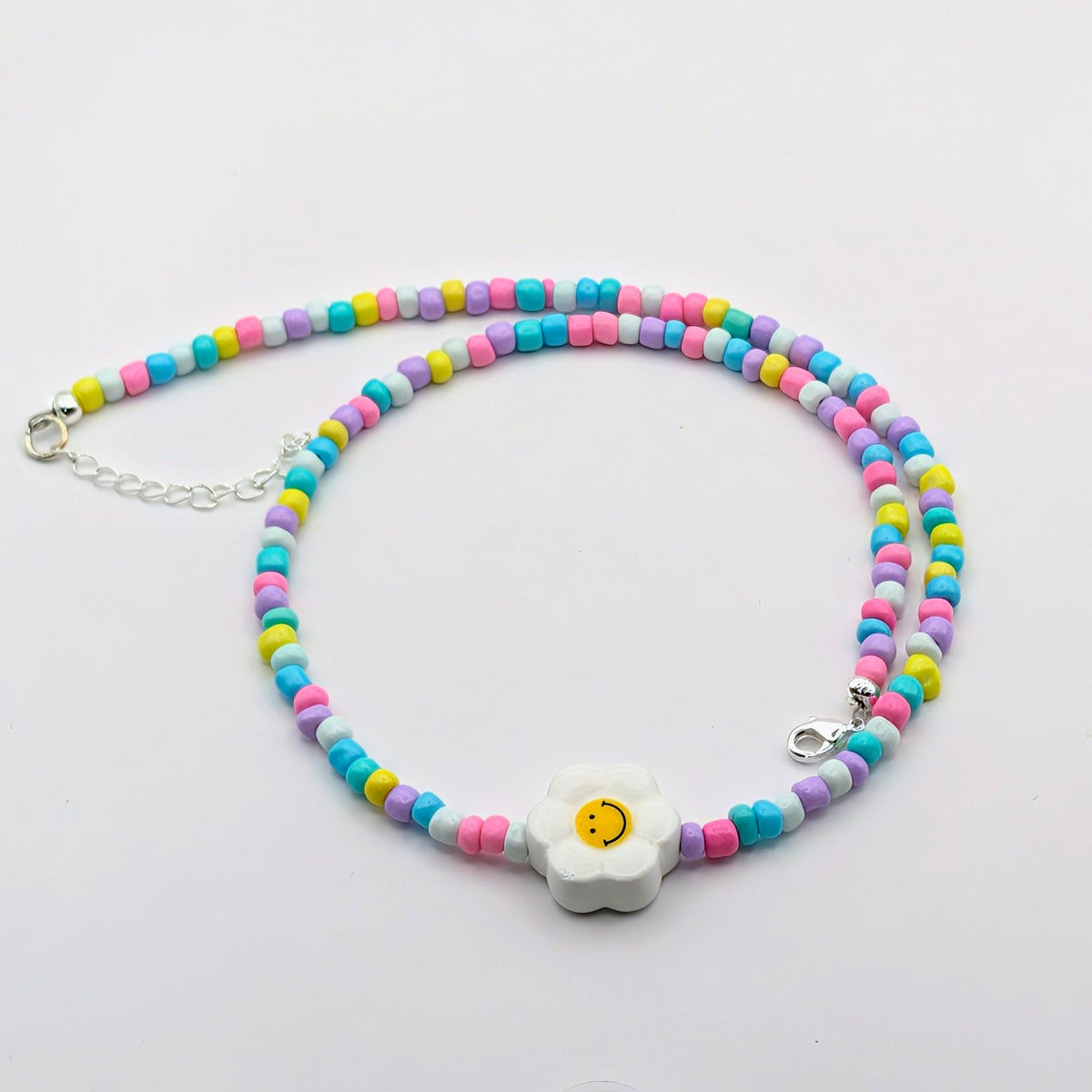 A pastel beaded necklace with a central white daisy charm featuring a yellow smiley face. The beads are arranged in two rows of alternating pastel pink, blue, yellow, and purple, with a silver lobster clasp and extender chain.