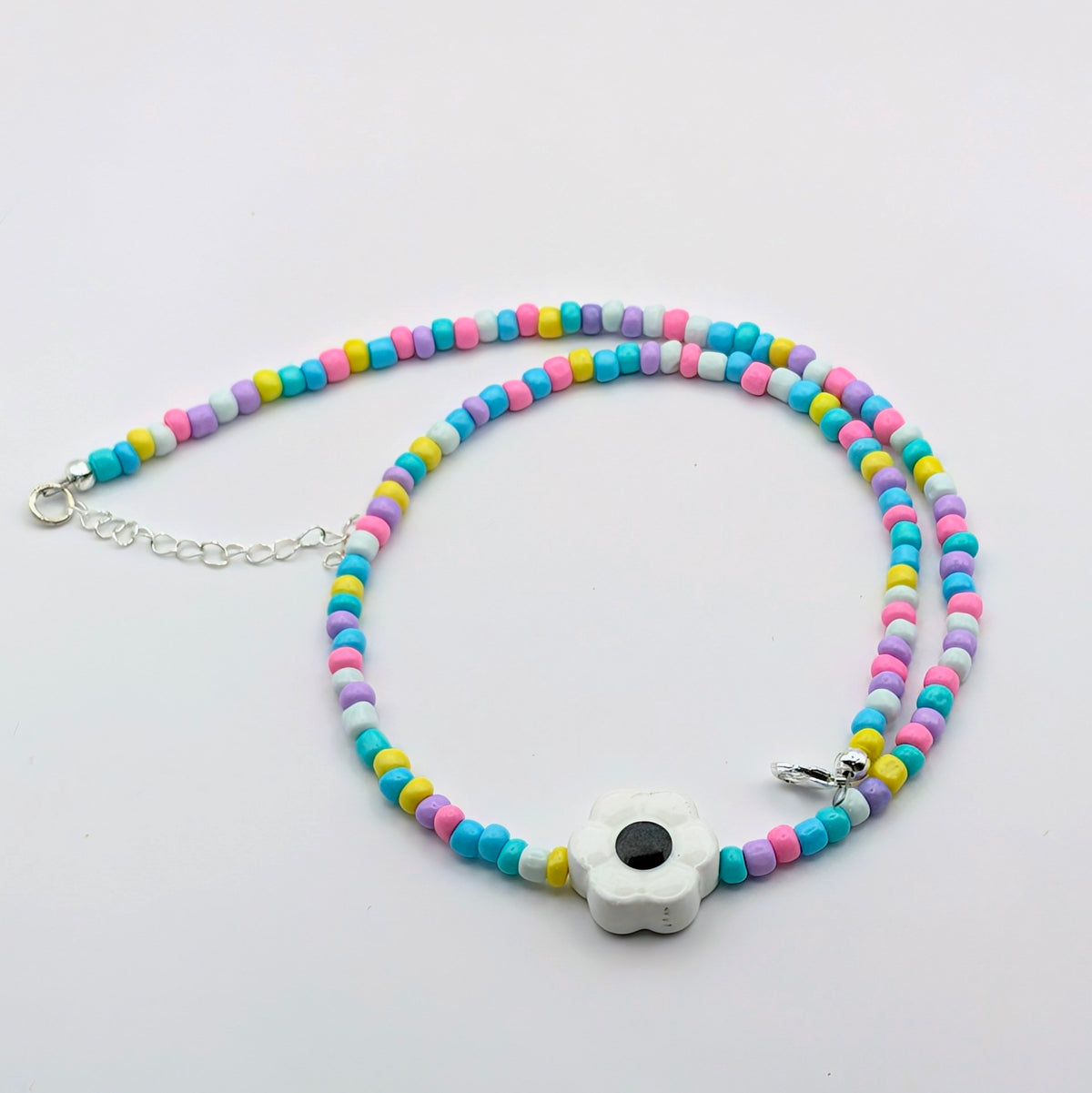 A beaded necklace with pastel blue, pink, yellow, and purple round beads, featuring a white flower-shaped charm with a black center and silver clasp, displayed on a clean white background.