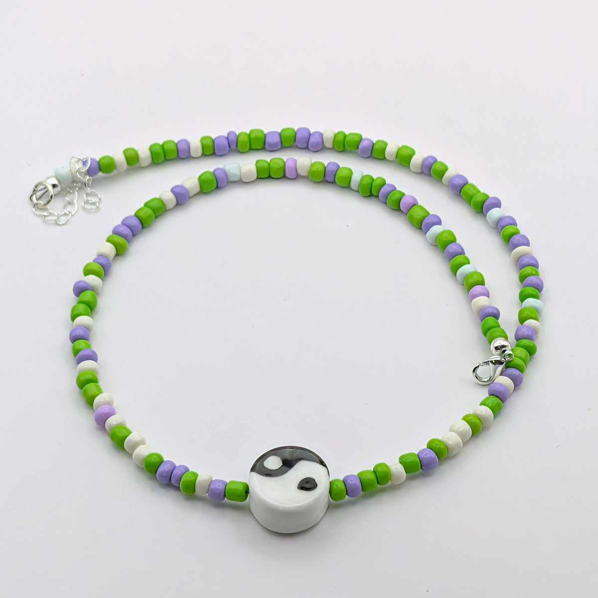 A beaded necklace with green, purple, and white cylindrical beads, featuring a white and black yin-yang pendant at the center and a silver lobster clasp.
