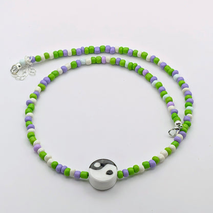 A beaded necklace with green, purple, and white cylindrical beads, featuring a white and black yin-yang pendant at the center and a silver lobster clasp.